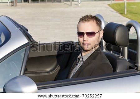 Young man in a dark suit and sunglasses, driving in his open sports car and is very relaxed.
