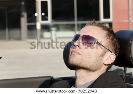 Young man in a dark suit and sunglasses, driving in his open sports car and is very relaxed.