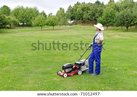 Adult man (gardener) in the blue overalls and straw hat with the lawn mower on a large lot