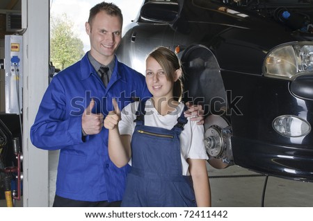 Mechanic in blue overalls in front of a car with the hood open smiles, pleased at the camera and makes the \'thumbs up\' gesture