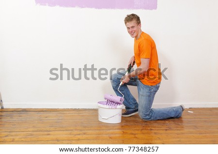 A young handsome man with blond hair in a orange T-shirt renovated his apartment. He strikes the wall with paint and a paint roller.