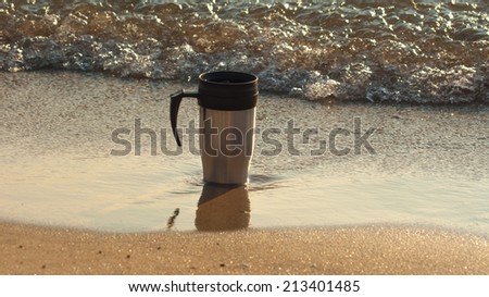 Heat protection-thermos coffee cup on the beach, close to the waves and nestled into the center of grainy sand