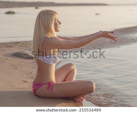 Slim blonde girl seat on the sand near the waves. Water streams between her fingers.