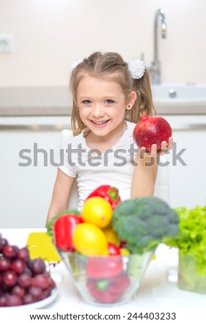 Lovely cute smiling girl shows a pomegranate, near is a bowl with vegetables - broccoli, paprika, lemons, dish with grapes and salad.