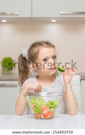 Little young cute sweet smiling girl eats a cucumber slice while make fresh salad
