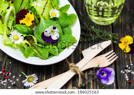 Salad leaves with wild flowers and herbs on a white plate on a vintage wooden surface, a glass of water, wooden cutlery, sea salt and pepper