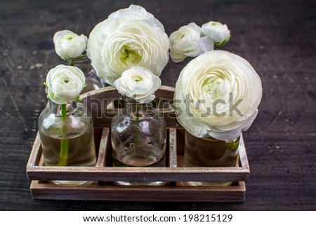 White ranunculus flowers in glass bottles in a wooden tray on a shabby surface of natural wood