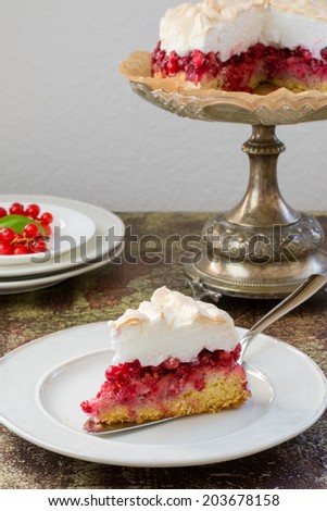 Red currant meringue tart on a vintage cake stander, a slice of tart on a cake server placed on a white plate and a stack of white dishes with fresh red currants on a vintage painted surface