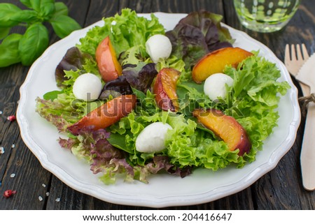 Close-up of assorted salad leaves, grilled peaches and mozzarella salad on a white vintage plate placed on a dark wooden surface; glass of water and fresh basil on the background