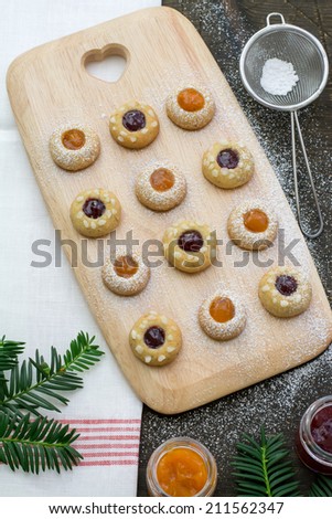 Thumbprint cookies with raspberry and apricot jams on a wooden serving board, glasses of jam and decorating sugar placed on a natural dark wood surface covered with the white red tea towel