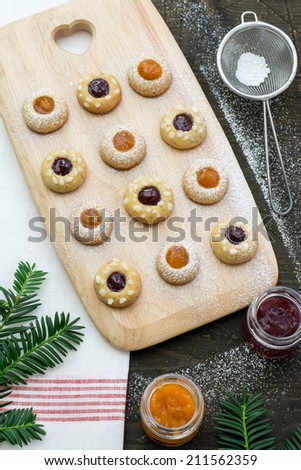 Thumbprint cookies with raspberry and apricot jams on a wooden serving board, glasses of jam and a sieve placed on a natural dark wood surface covered with the white red tea towel