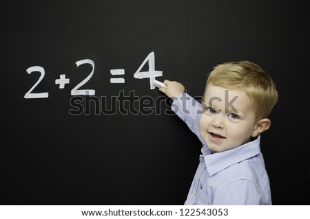 Smart young boy wearing a blue striped shirt stood writing a math sum on a blackboard