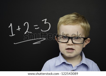 Smart young boy wearing a blue striped shirt and glasses stood in front of a blackboard with a drawn on chalk addition sum