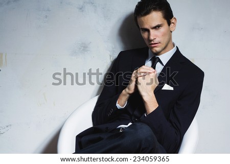Serious man in a black business suit sitting on a white chair on a white grungy background. Studio shoot