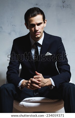 Serious man in a black business suit sitting on a white chair on a white grungy background. Studio shoot