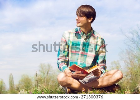 Young handsome man sitting cross-legged on green grass with book on blue sky background.