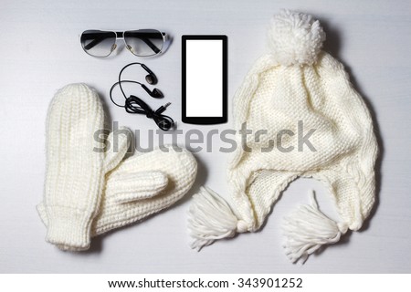 still life of winter accessories. white hat, mittens,phone, sunglasses and headphones lie on white wood background.