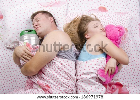 Young couple lying in bed facing away from each other. Husband sleeps with a barrel of beer, a wife with a soft toy