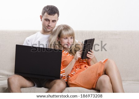 A boy and a girl sitting on the couch. A guy sitting with a laptop, a girl with a tablet. The girl clung to the guy. Both smiling,