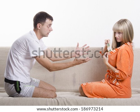 Young couple sitting on the couch, facing each other. Wife hides from her husband of bills, a Bathrobe, a husband and stretches her hands to her imploring her to give him money.