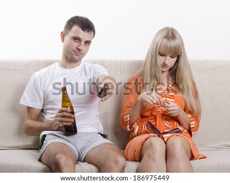 Couple sits on the couch. He watches TV, remote switch channels, drink beer, it handles nails nail file.