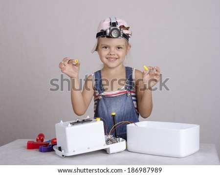 Girl playing in the repairer and repairs toy microwave. In one hand it a cog in another nut. Girl with fun and pleasure looks into the frame, and raised her hands. child\'s head wearing a flashlight