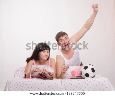 Young girl and a guy in bed. Beautiful girl has a manicure, man watches football on TV.