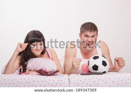 Young girl and a guy in bed. Beautiful girl has a manicure, man watches football on TV.