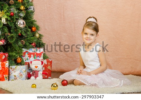 Five-year girl in a New Year\'s dress sitting on a mat at the Christmas tree