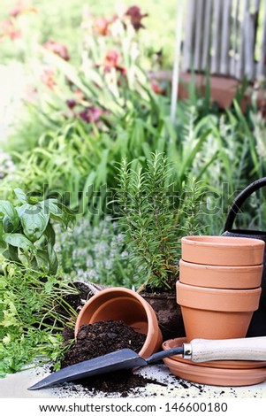 Rustic table with terracotta pots, potting soil, trowel and herbs in front of a beautiful garden surrounding a rustic porch.