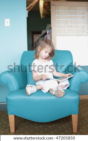 Little girl sitting in a colorful chair looks at or is reading a book.