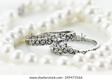 Silver diamond engagement ring and wedding band lying inside of a beautiful pearl necklace. Extreme shallow depth of field with selective focus on rings.