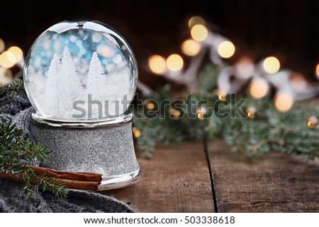 Rustic image of a Christmas snow globe surrounded by pine branches, cinnamon sticks and a warm gray scarf. Shallow depth of field with selective focus on snowglobe.