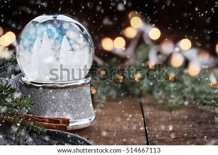 Rustic image of a snow globe surrounded by pine branches, cinnamon sticks and a warm gray scarf with falling snow. Shallow depth of field with selective focus on snowglobe.