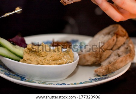 White Plate of hummus cucumber, salad leaves and bread with hand holding a slice of bread and knife