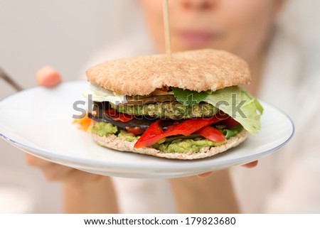 Girl holding delicious vegan burger with pea patty, grilled pear, grilled red peppers, shredded carrot, iceberg salad, ketchup, pickles and onions on white plate in hand