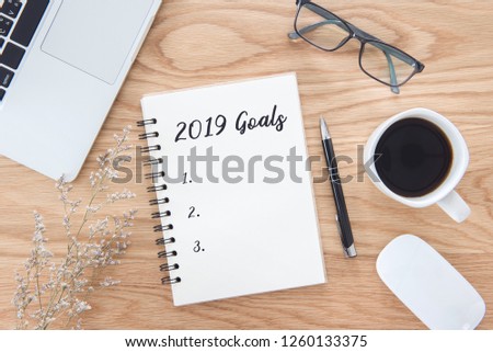 New year 2019 goals list in notebook with laptop, mouse, pen, glasses, green leaf, and coffee cup on wooden table background.