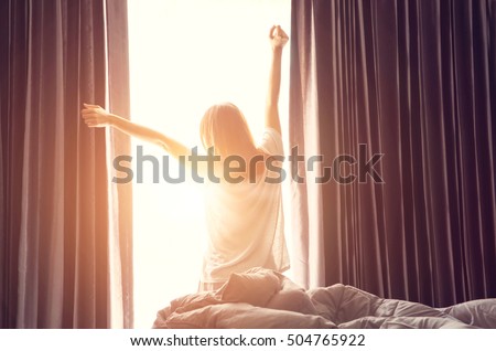 Woman standing near the window while stretching near bed after waking up with sunrise at morning, back view