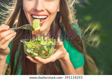 Woman eating healthy salad from plastic container over green grass with flying hair on a sunny spring day. backlight