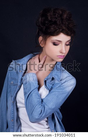 Beautiful young girl in jeans coat looking down, touching her neck. Black background.