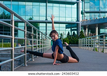 Girl sitting on the splits, with one foot on a metal fence. Beautiful girl performs acrobatics. ballerina in casual clothes in the street. Street dance
