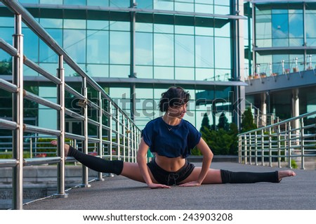 Girl sitting on the splits, with one foot on a metal fence. Beautiful girl performs acrobatics. ballerina in casual clothes in the street. Street dance