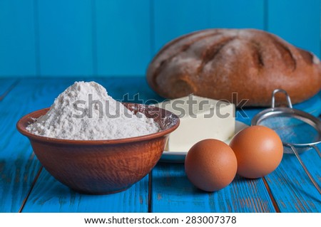 Baking bread in rural kitchen - dough recipe ingredients eggs, flour, butter, sugar and whisk, screen on vintage blue wood table from above.