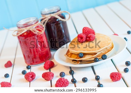 Stack of wheat golden pancakes or pancake cake with freshly picked raspberries on a dessert plate, glass mason jar full off blueberry and raspberry jam. Cooking background, unique perspective.