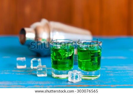 Closeup of two green absinthe alcohol shots with ice cubes and shaker on a light wooden background
