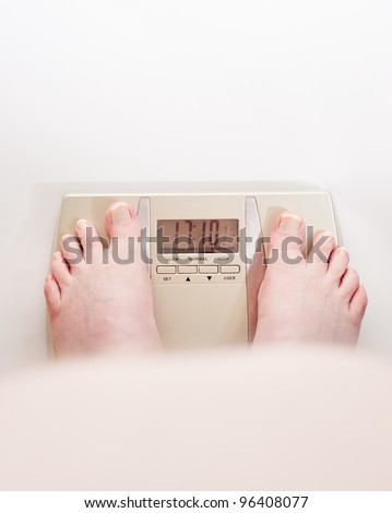 Obese man looking over his fat stomach at his feet on the weighing scales.