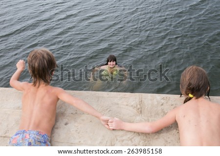 Fraternal twins tan holding hands on wooden deck at the lake on hot summer day. Mother swimming on the background. Family having fun during vacation.