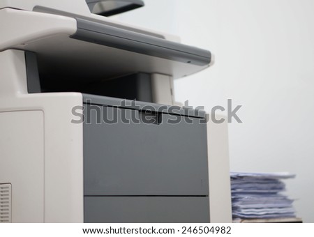 White copiers located in the office, side of the machine has a pile of papers.