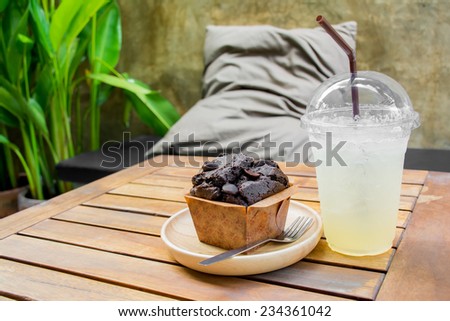 Chocolate muffin and lemonade on wooden table with cushion pillow background.