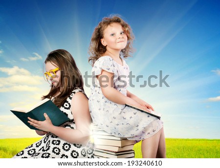 Two pretty young sisters reading books, one of them looking at camera. Blue sky.